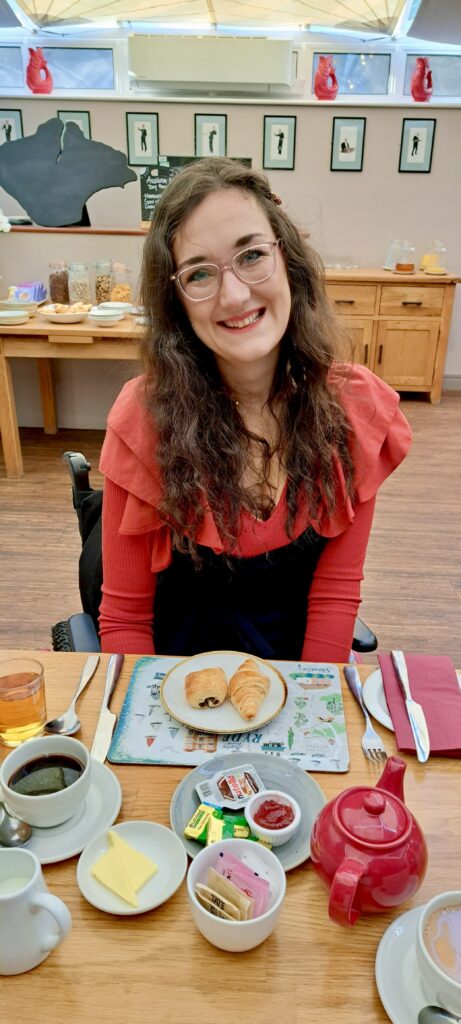 Georgina, a white brunette female who uses a wheelchair, wears an orange ruffled shirt and a black jumpsuit. She is at the breakfast dining table